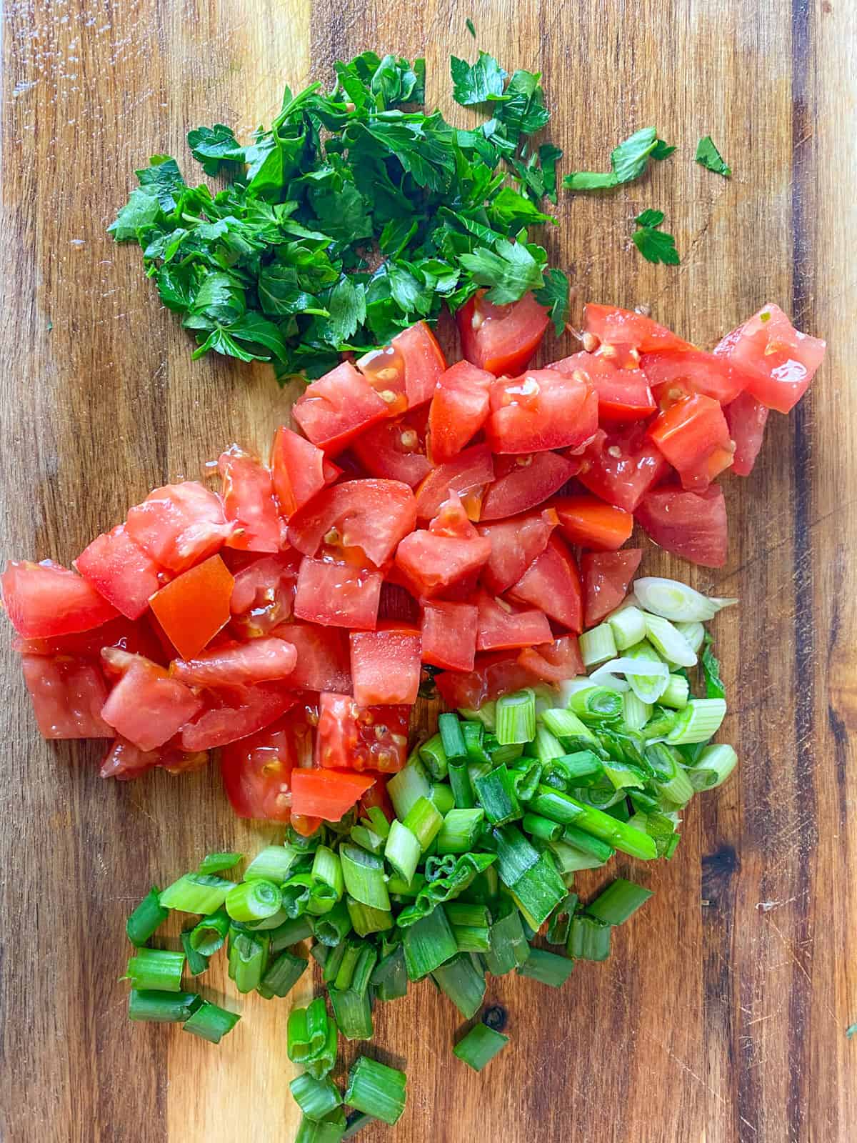 Chopped parsley, chopped tomatoes and sliced green onions on a wooden cutting board.