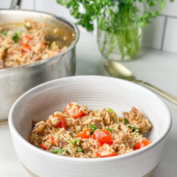 Spicy sausage rice in a white bowl with a tan bottom. Contents in bowl are light brown in color and topped with bright red tomatoes and green onions that have been lightly cooked. The background shows the pot of rice next to a gold spoon and fresh green parsley in a mason jar.