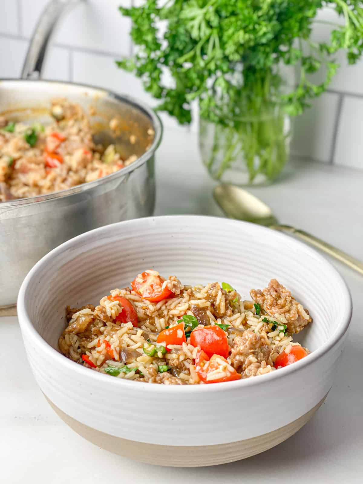 Spicy sausage rice in a white bowl with a tan bottom. Contents in bowl are light brown in color and topped with bright red tomatoes and green onions that have been lightly cooked. The background shows the pot of rice next to a gold spoon and fresh green parsley in a mason jar.