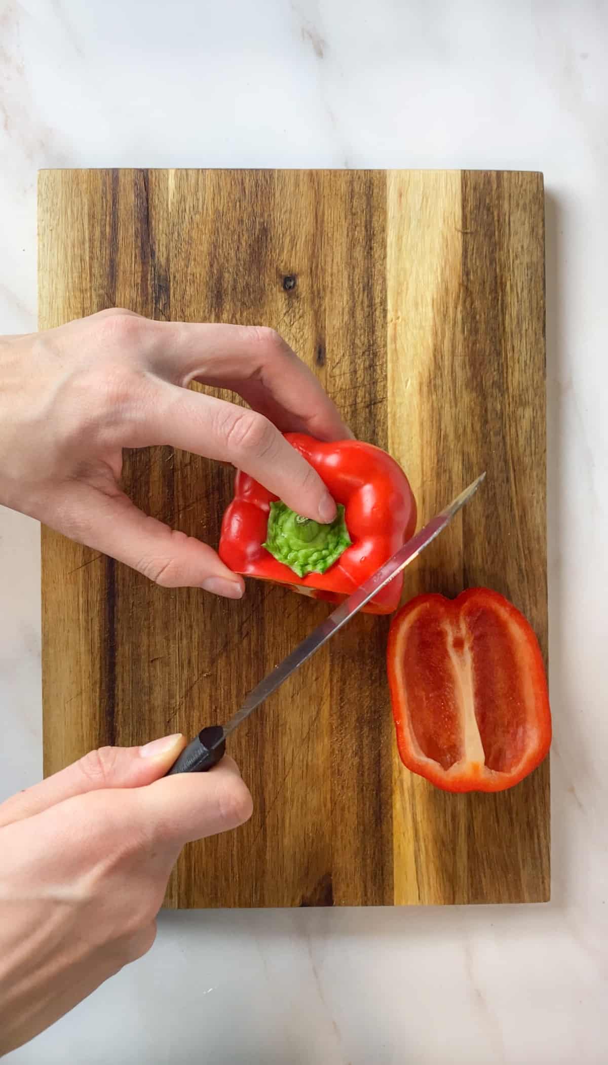 Slicing the sides off of a red bell pepper with a knife on a wooden cutting board.