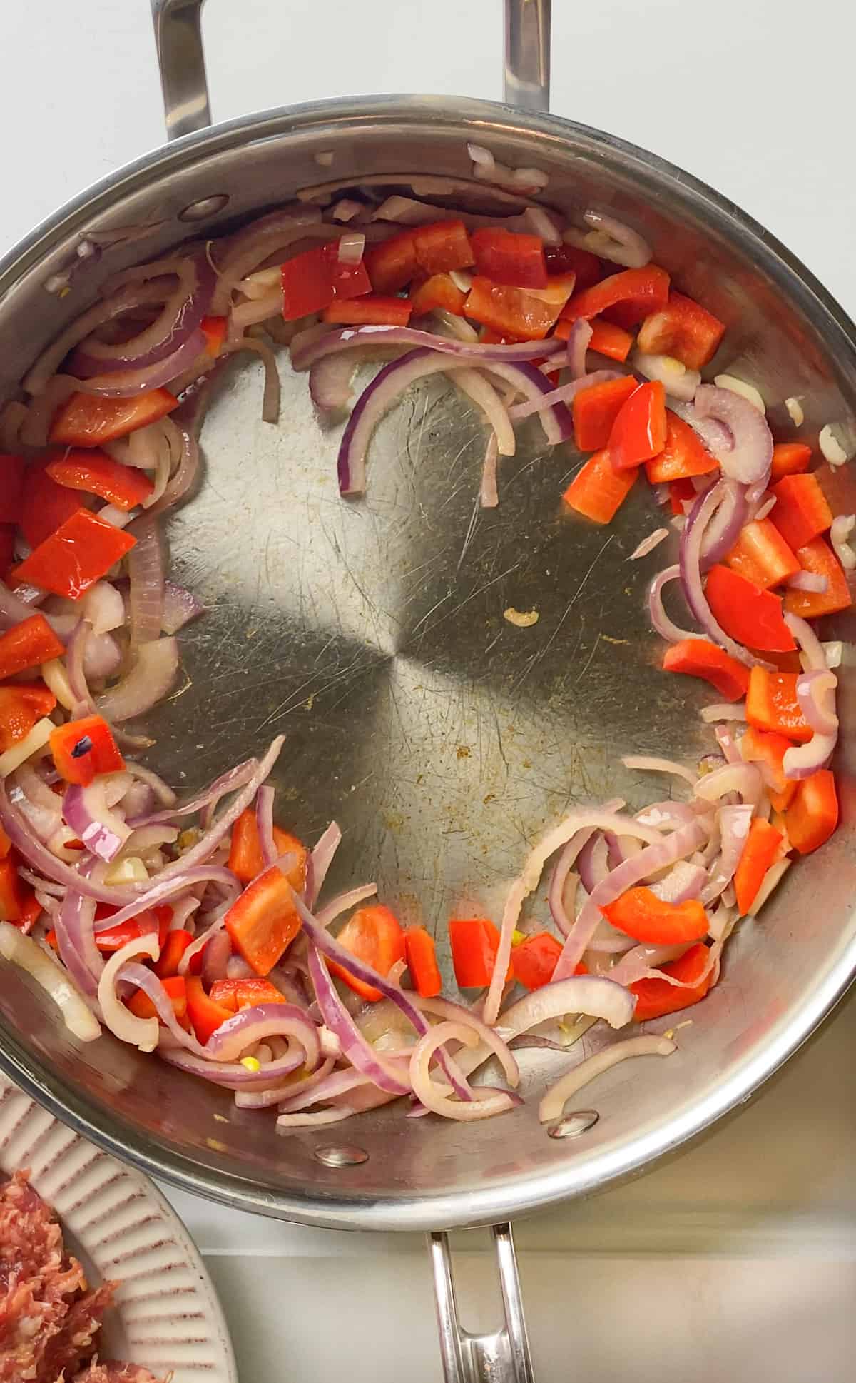 Sautéd red onions, red bell peppers and garlic pushed to the sides of the pan, making a well in the center. Sausage on a plate is in the bottom left corner of the image.