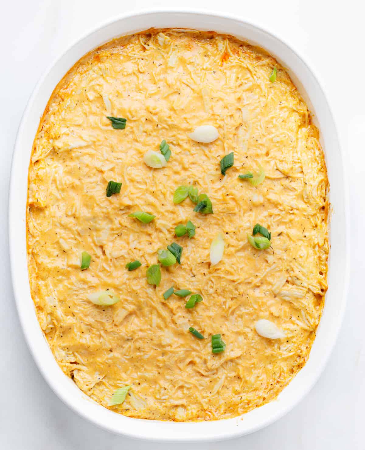 Overhead view of baked dairy-free buffalo chicken dip in a white casserole dish, topped with green onions on a white background.