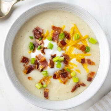 White bowl of potato soup on a white surface with a gold spoon in the top left corner. Soup is topped with crisp bacon bits, vegan cheddar shreds, green onions and a sour cream alternative.