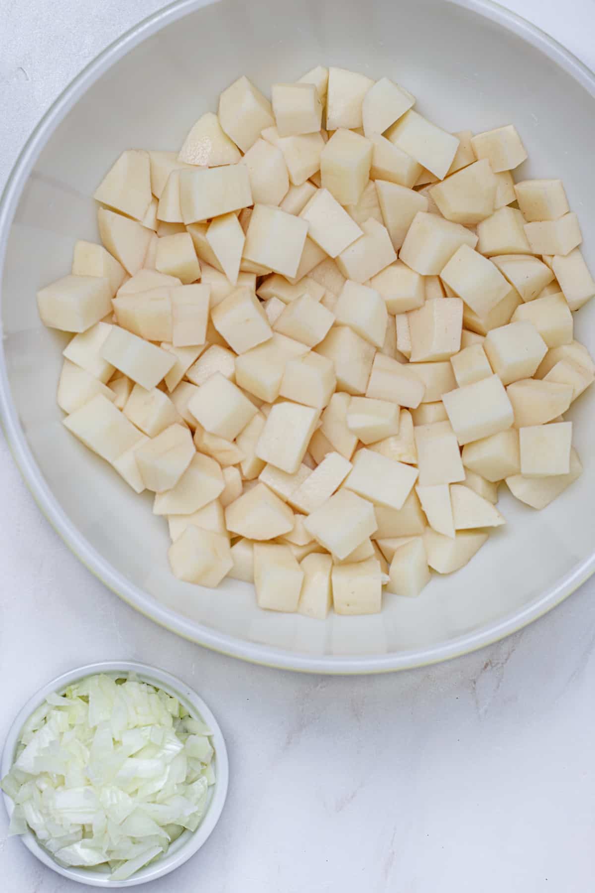 Large bowl of peeled and diced russet potatoes with a small bowl of diced onions in the bottom left corner.