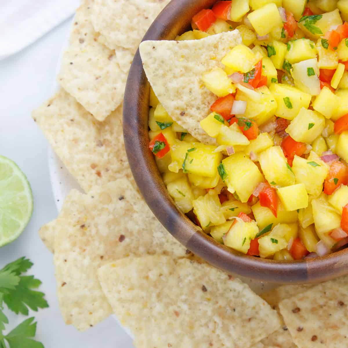 Pineapple Mango Habanero Salsa in a wooden bowl on top of a white plate with tortilla chips. Salsa shows yellow mangos and pineapples, with red bits of bell pepper and small bits of red onion and cilantro. The left of the photo shows the inside of half a lime with green cilantro next to it.