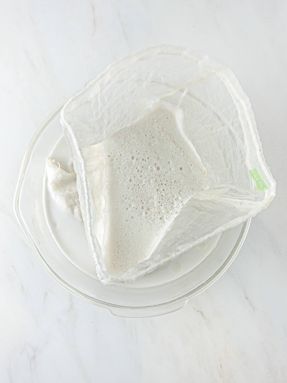 Homemade almond milk being strained through a nut milk bag in a large glass bowl.