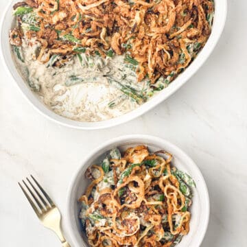 Overhead shot of dairy-free green bean casserole in a white bowl with a gold fork to it's left, and a white casserole dish filled with green bean casserole behind it.