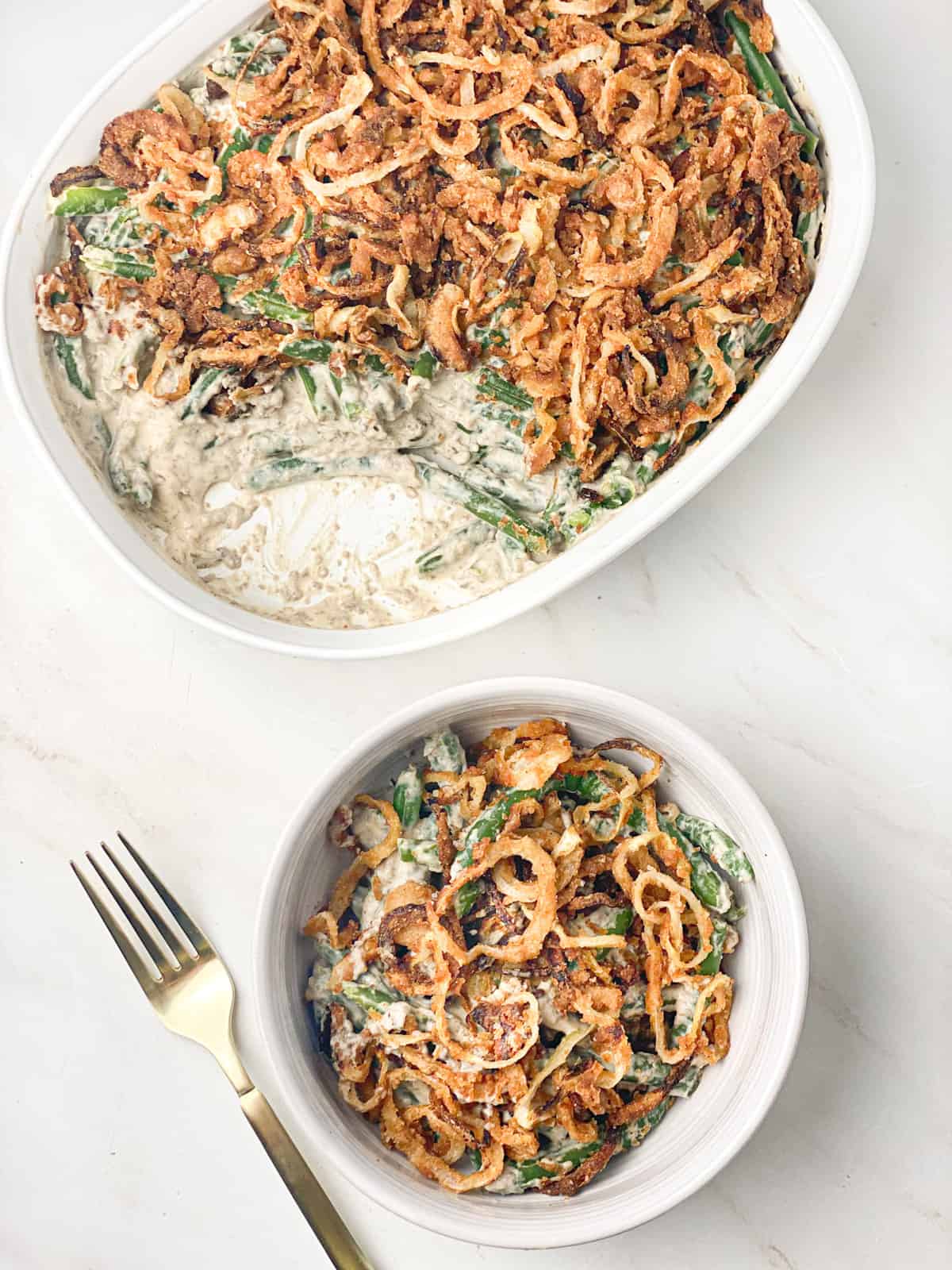 Overhead shot of dairy-free green bean casserole in a white bowl with a gold fork to it's left, and a white casserole dish filled with green bean casserole behind it.