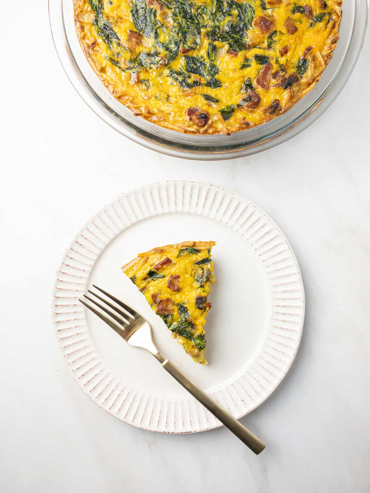 A slice of dairy-free gluten-free quiche on a white plate with a gold fork with the pie dish behind it.