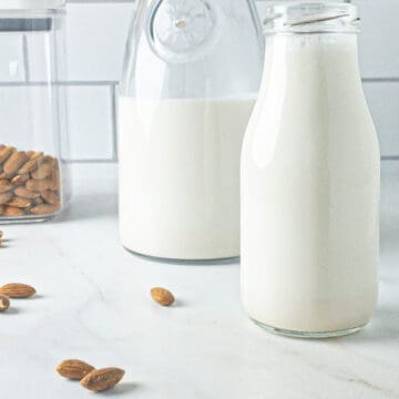 Homemade almond milk in a milk bottle with a carafe of almond milk behind it, and a container of almonds beside it.