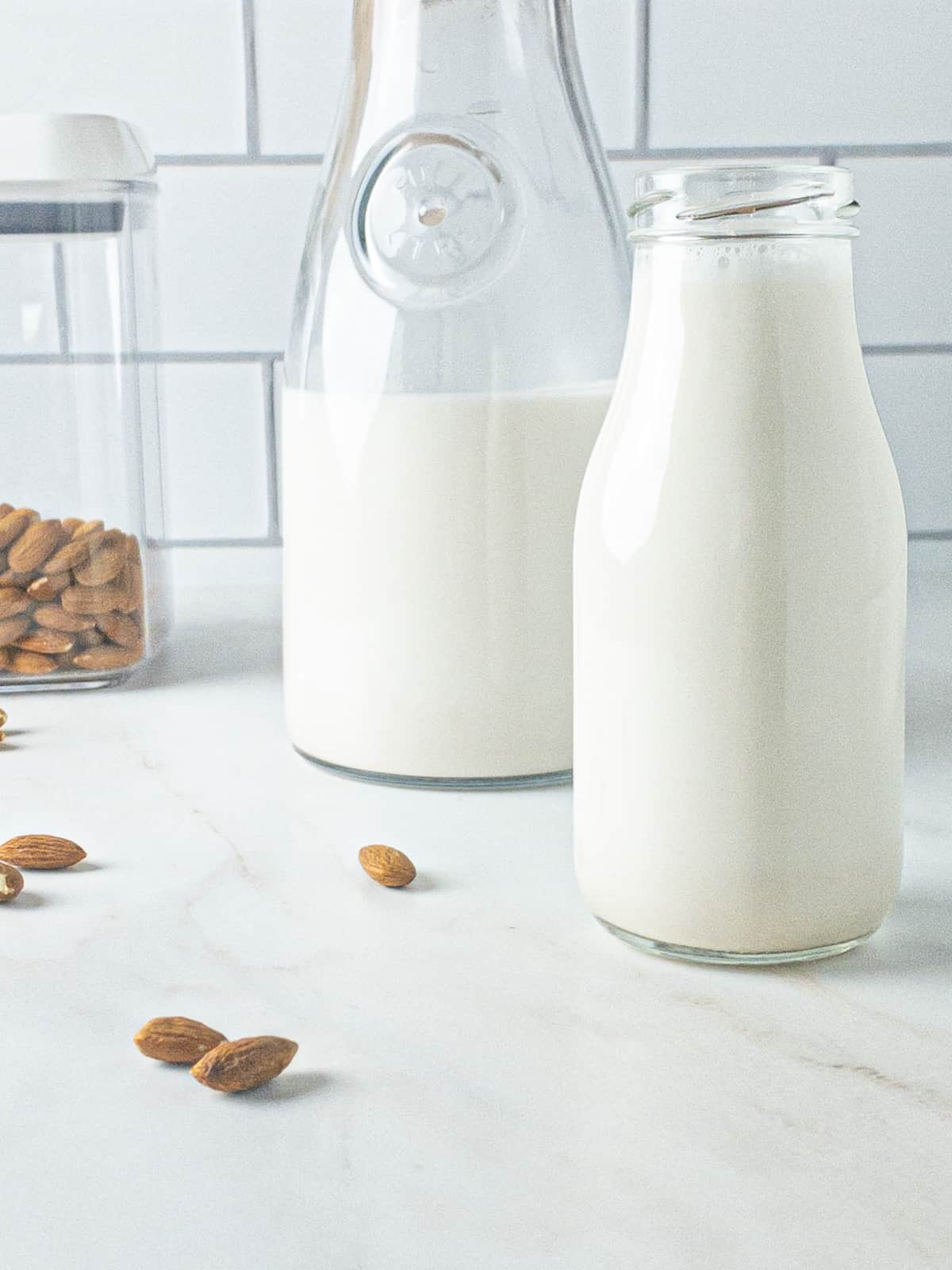 Homemade almond milk in a milk bottle with a carafe of almond milk behind it, and a container of almonds beside it.