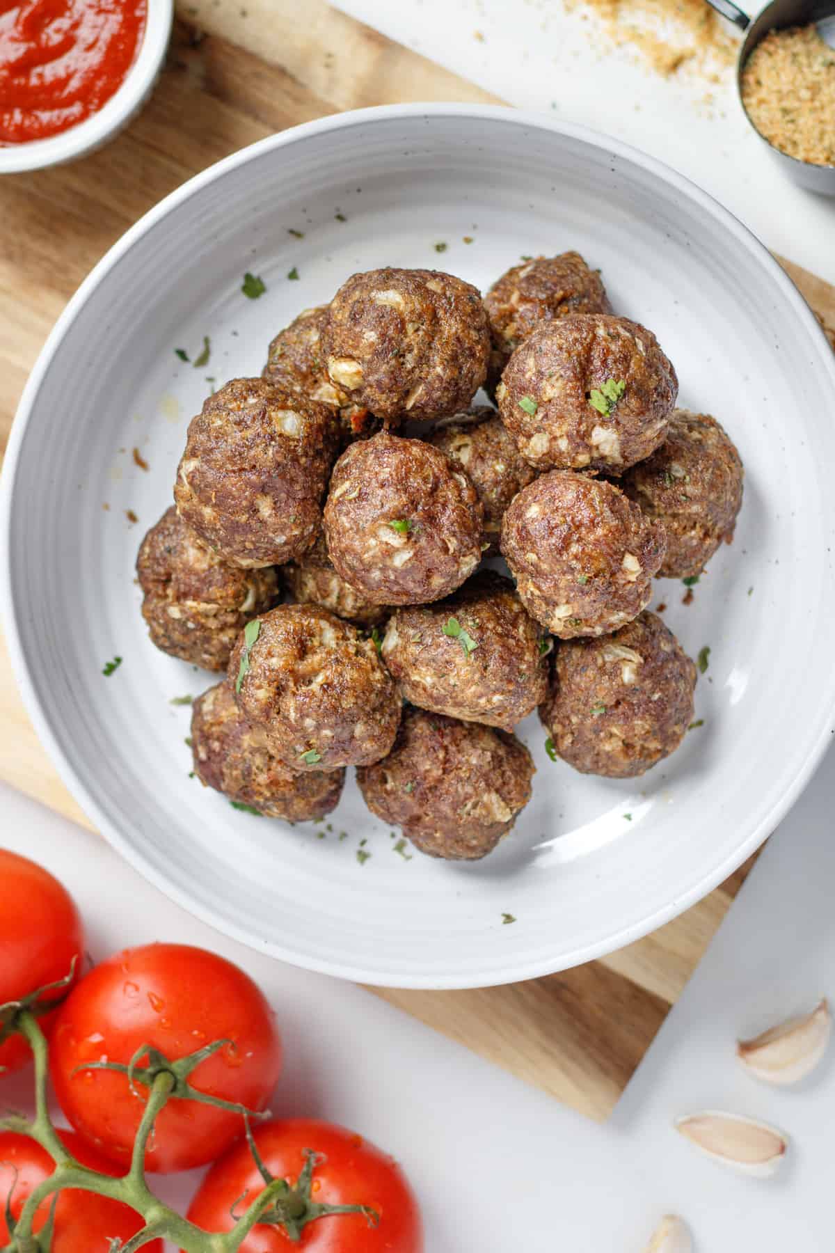 Wagyu meatballs in a white bowl on top of a wooden cutting board. Surrounding the bowl there are red tomatoes on the vine, garlic cloves, marinara sauce and spilled bread crumbs in a black measuring cup.