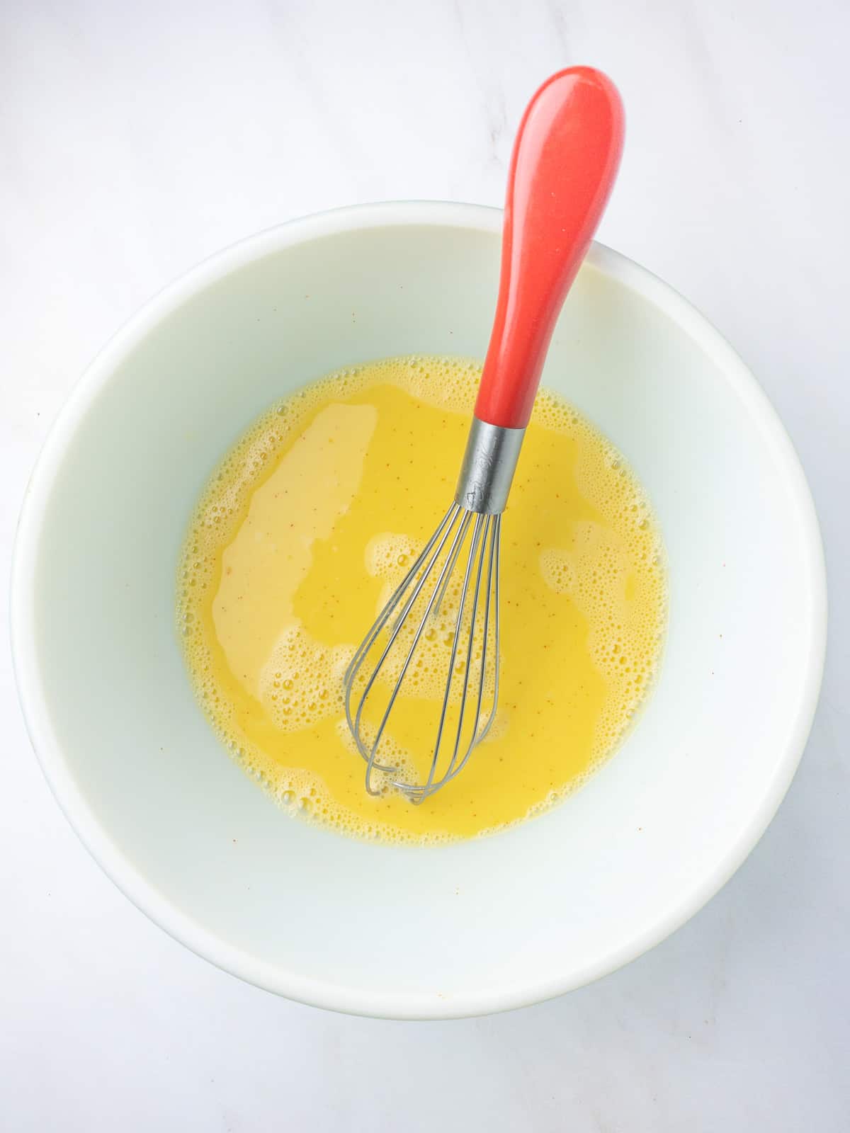 Whisked eggs in a bowl with a red handled whisk.