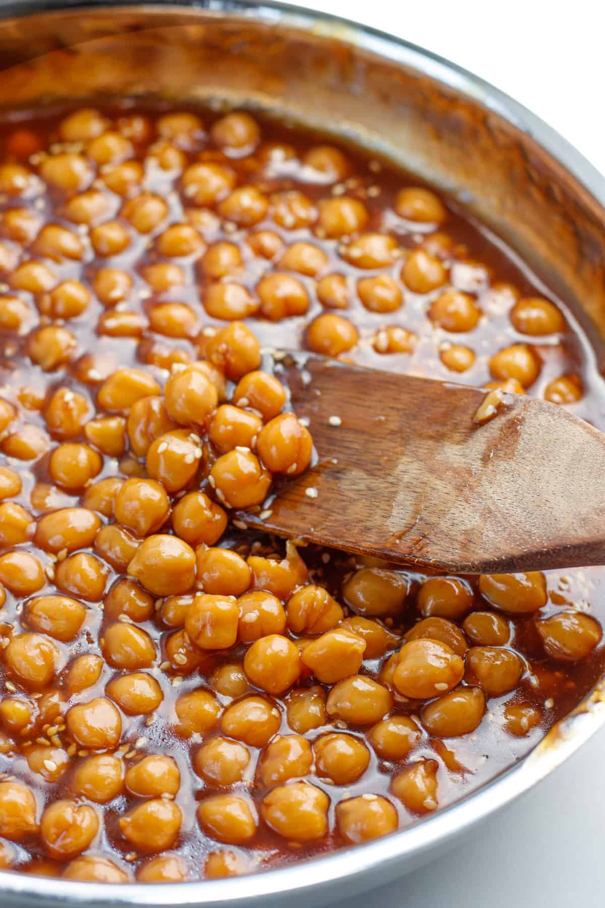 Sticky sesame chickpeas in a stainless steel pan with a wooden spoon in the pan.
